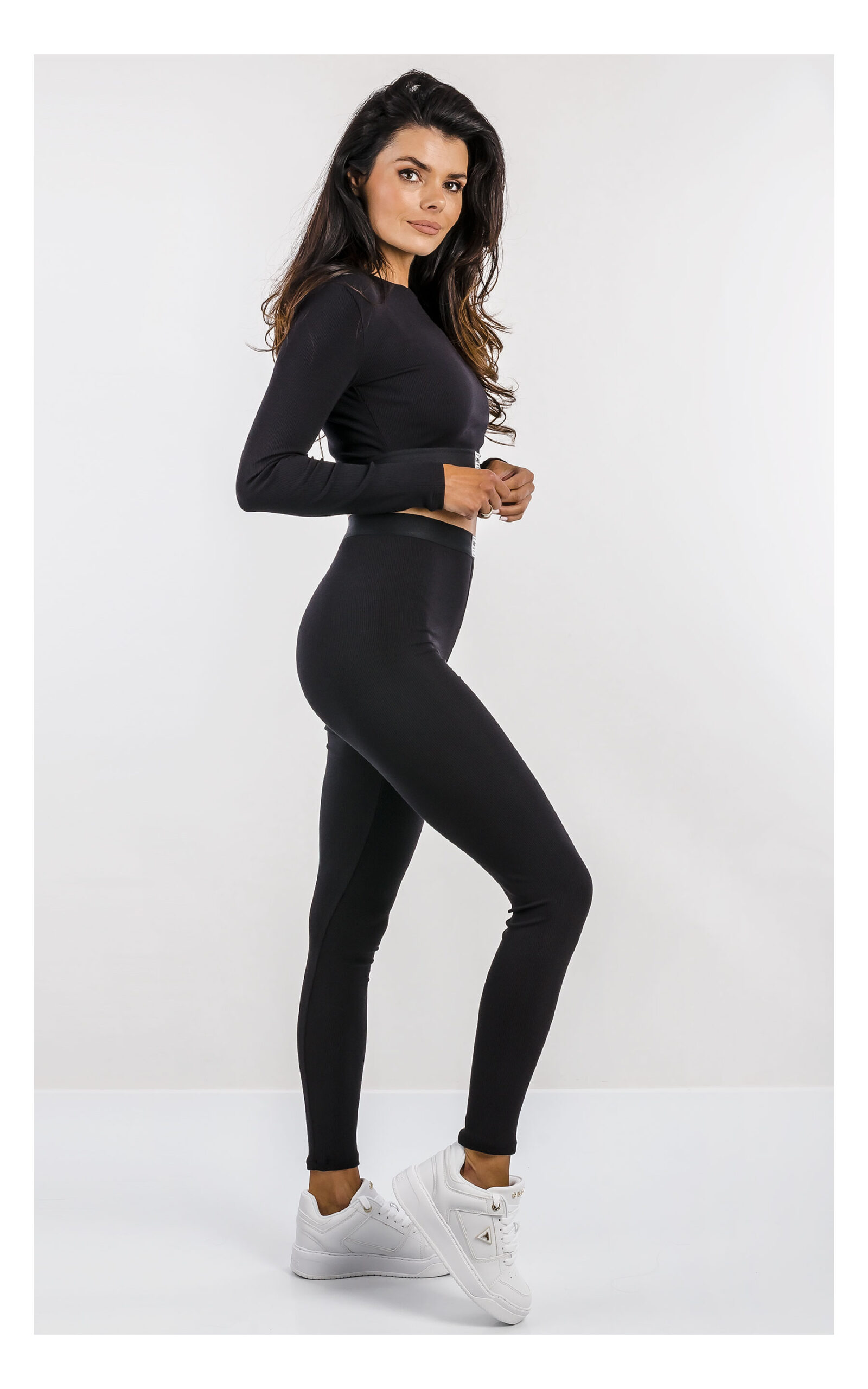 czarny crop top z długim rękawem, dopasowany z logowaną naszywką. Stylizacja z legginsami do kompletu. widok na modelce z profilu