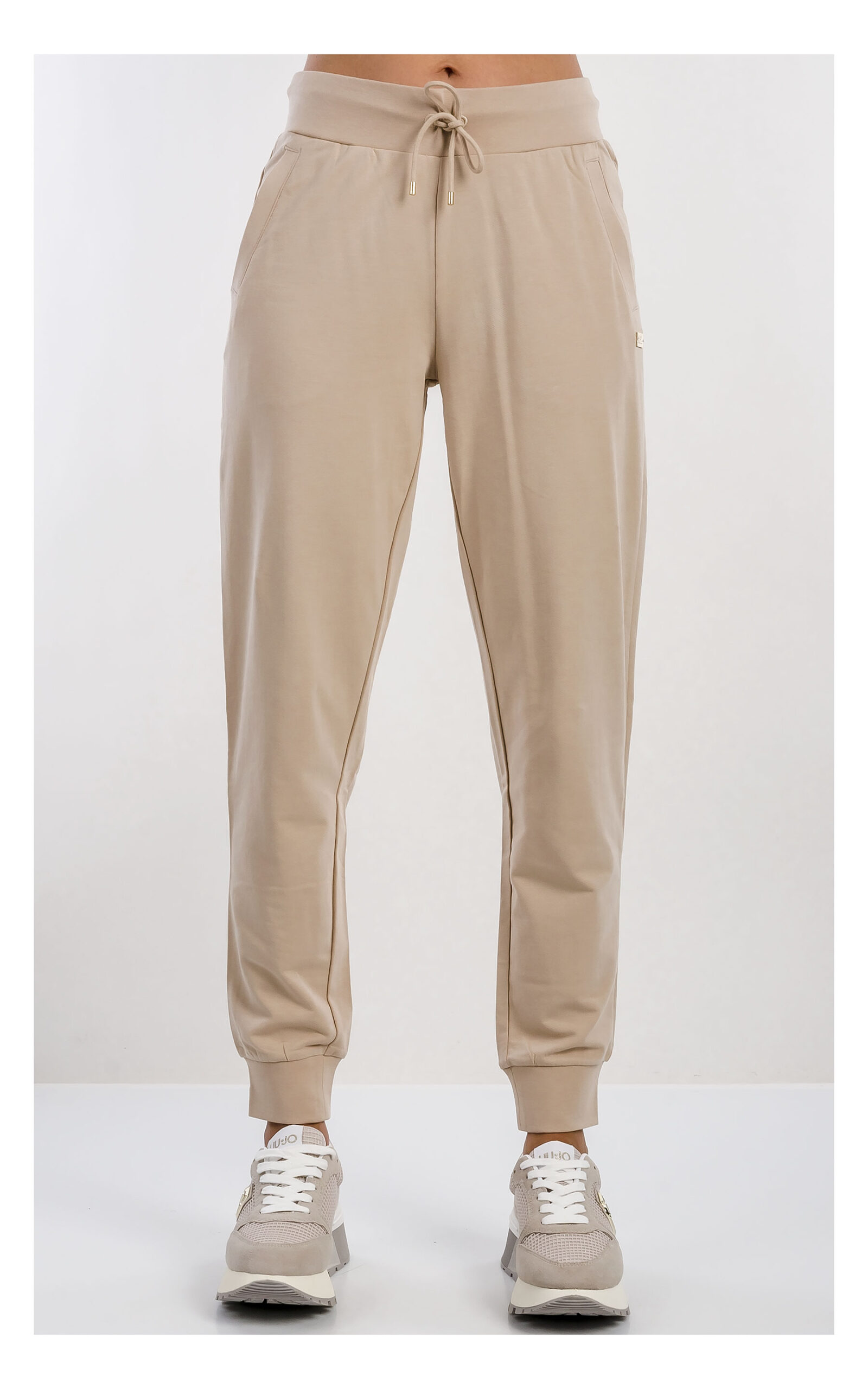 Beżowy komplet damski EA7 Emporio Armani z cienkiej dzianiny, składający się z beżowych spodni dresowych z elastycznym pasem i ściągaczami oraz rozpinanej bluzy z kapturem, z logo EA7 na piersi, prezentowany na jasnym tle, zbliżenie na spodnie z kompletu od przodu