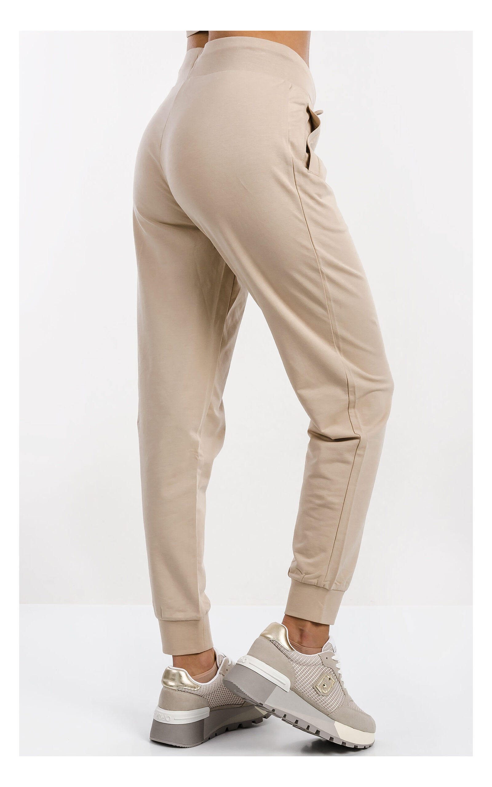 Beżowy komplet damski EA7 Emporio Armani z cienkiej dzianiny, składający się z beżowych spodni dresowych z elastycznym pasem i ściągaczami oraz rozpinanej bluzy z kapturem, z logo EA7 na piersi, prezentowany na jasnym tle, zbliżenie na spodnie z kompletu od tyłu