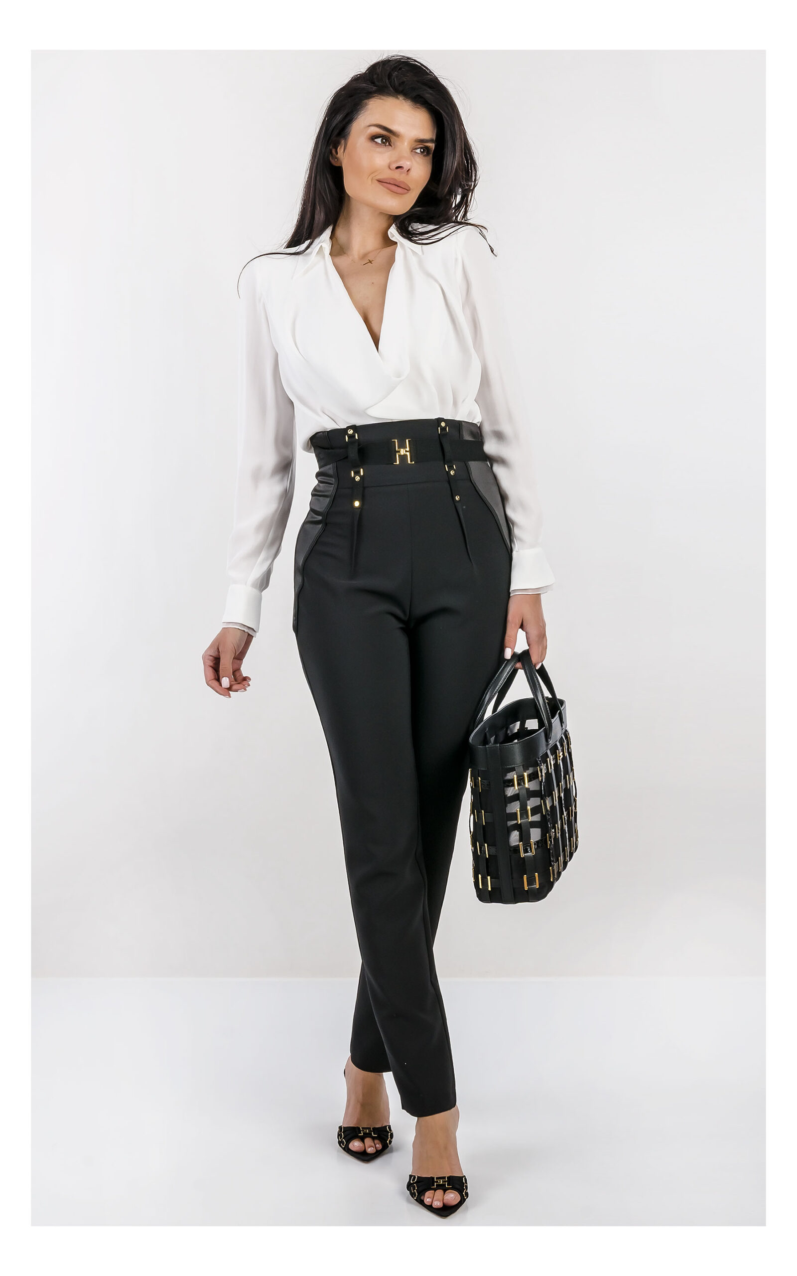 Na zdjęciu znajduje się modelka na jasnym, studyjnym tle, ubrana w elegancką stylizację. Ma na sobie białe body z długim rękawem i głębokim dekoltem oraz dopasowane, wysokie spodnie z ozdobnym pasem i złotymi detalami. W dłoni trzyma czarną torebkę Elisabetta Franchi z dekoracyjnymi elementami w kolorze złota. Stylizację uzupełniają czarne klapki na obcasie z metalicznymi zdobieniami. Cała stylizacja jest od marki Elisabetta Franchi.