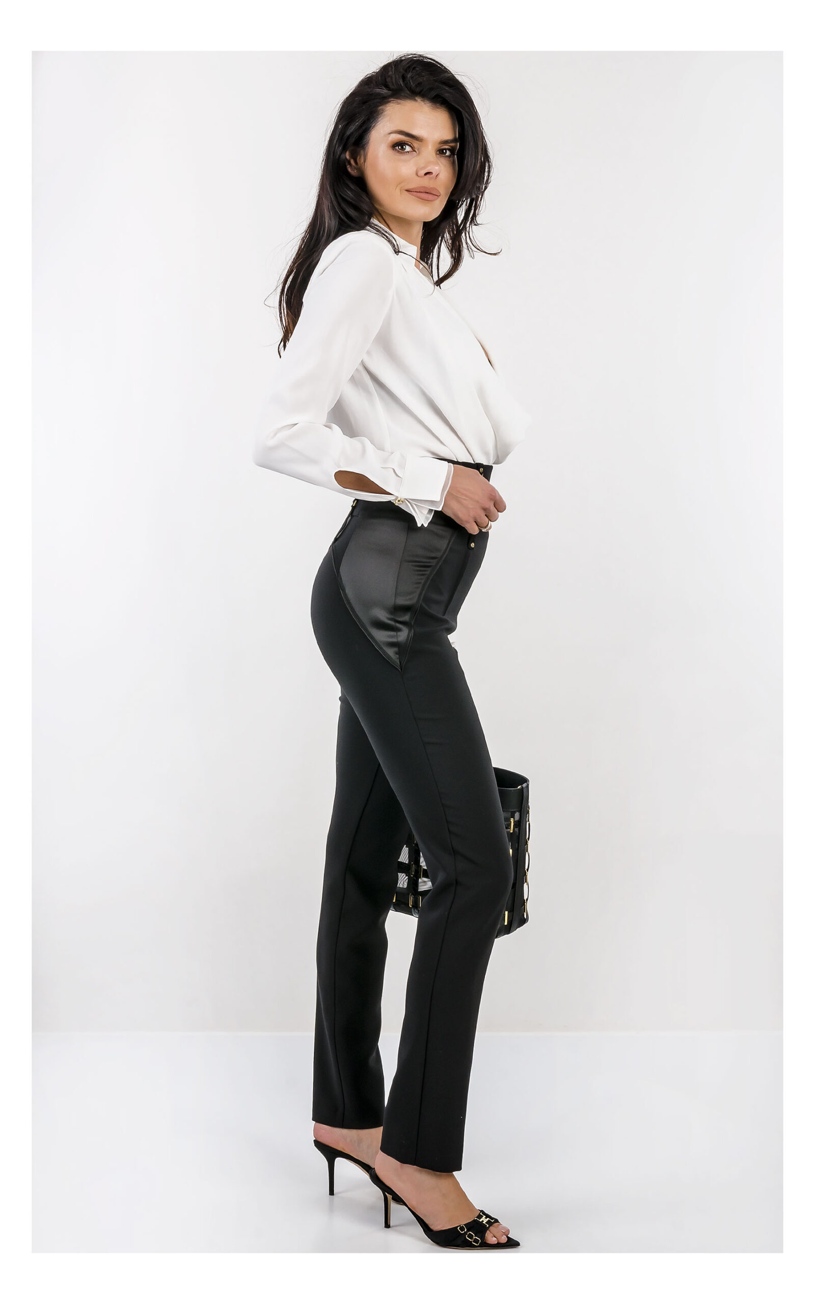 Na zdjęciu znajduje się modelka na jasnym, studyjnym tle, ubrana w elegancką stylizację. Ma na sobie białe body z długim rękawem i głębokim dekoltem oraz dopasowane, wysokie spodnie z ozdobnym pasem i złotymi detalami. W dłoni trzyma czarną torebkę Elisabetta Franchi z dekoracyjnymi elementami w kolorze złota. Stylizację uzupełniają czarne klapki na obcasie z metalicznymi zdobieniami. Cała stylizacja jest od marki Elisabetta Franchi.