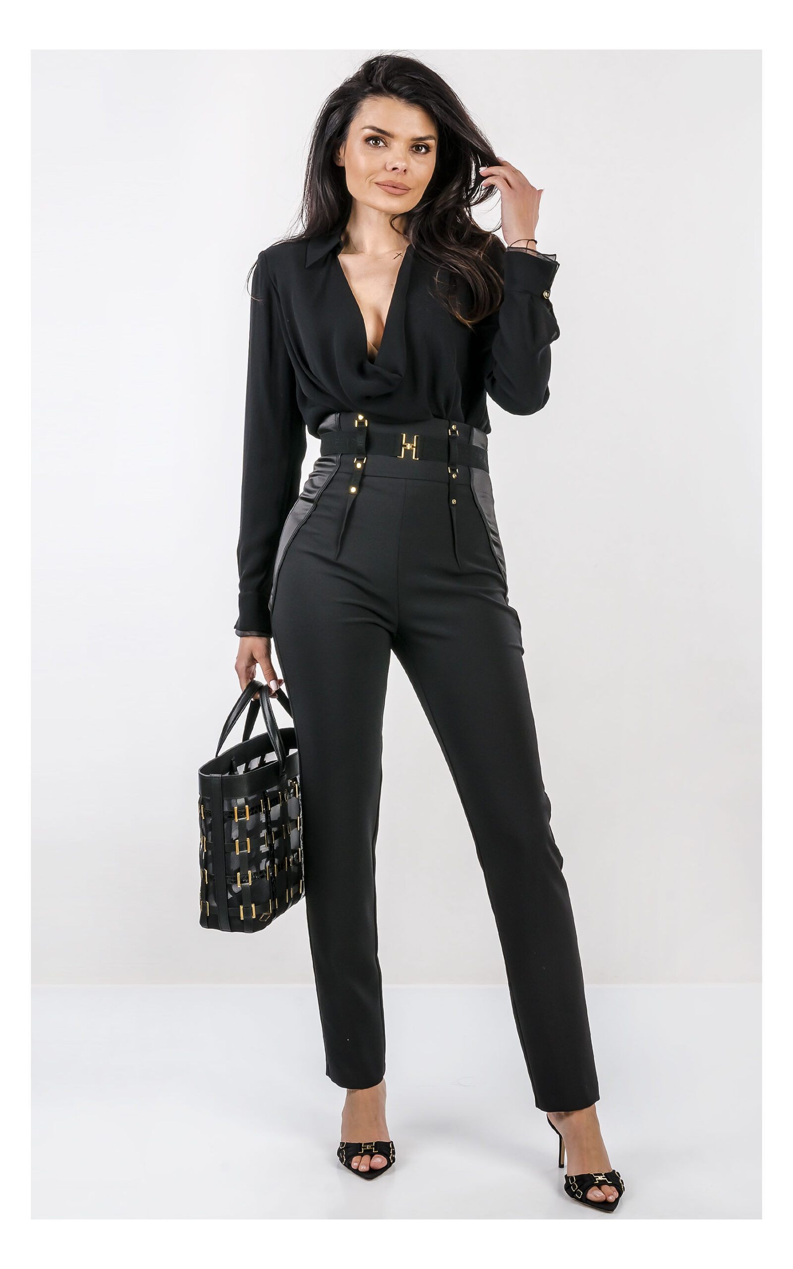Na zdjęciu znajduje się modelka na jasnym, studyjnym tle, ubrana w elegancką stylizację w całości w kolorze czarnym. Ma na sobie czarne body z długim rękawem i głębokim dekoltem oraz dopasowane, wysokie spodnie z ozdobnym pasem i złotymi detalami. W dłoni trzyma czarną torebkę Elisabetta Franchi z dekoracyjnymi elementami w kolorze złota. Stylizację uzupełniają czarne klapki na obcasie z metalicznymi zdobieniami. Cała stylizacja jest od marki Elisabetta Franchi.