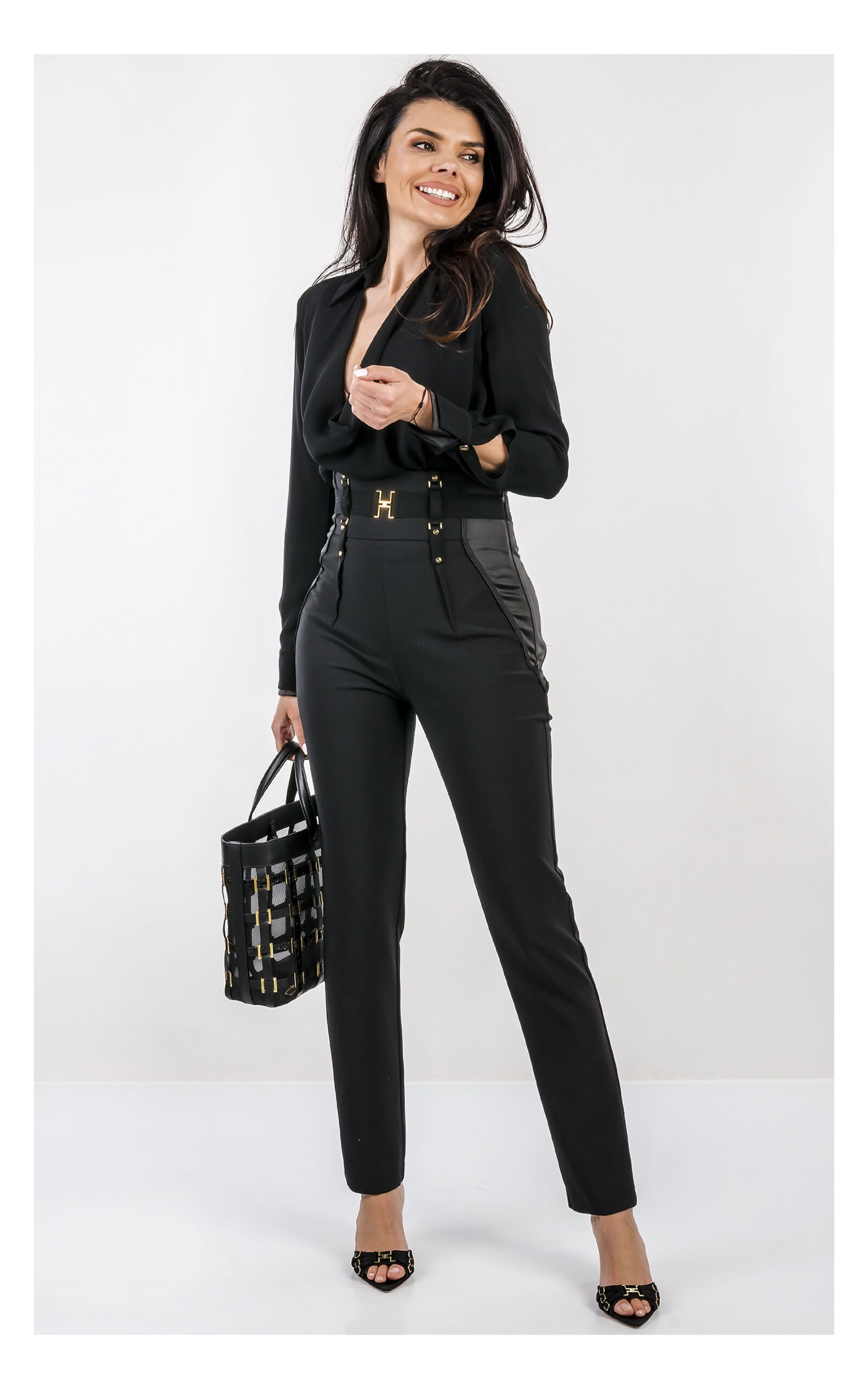 Na zdjęciu znajduje się modelka na jasnym, studyjnym tle, ubrana w elegancką stylizację w całości w kolorze czarnym. Ma na sobie czarne body z długim rękawem i głębokim dekoltem oraz dopasowane, wysokie spodnie z ozdobnym pasem i złotymi detalami. W dłoni trzyma czarną torebkę Elisabetta Franchi z dekoracyjnymi elementami w kolorze złota. Stylizację uzupełniają czarne klapki na obcasie z metalicznymi zdobieniami. Cała stylizacja jest od marki Elisabetta Franchi.
