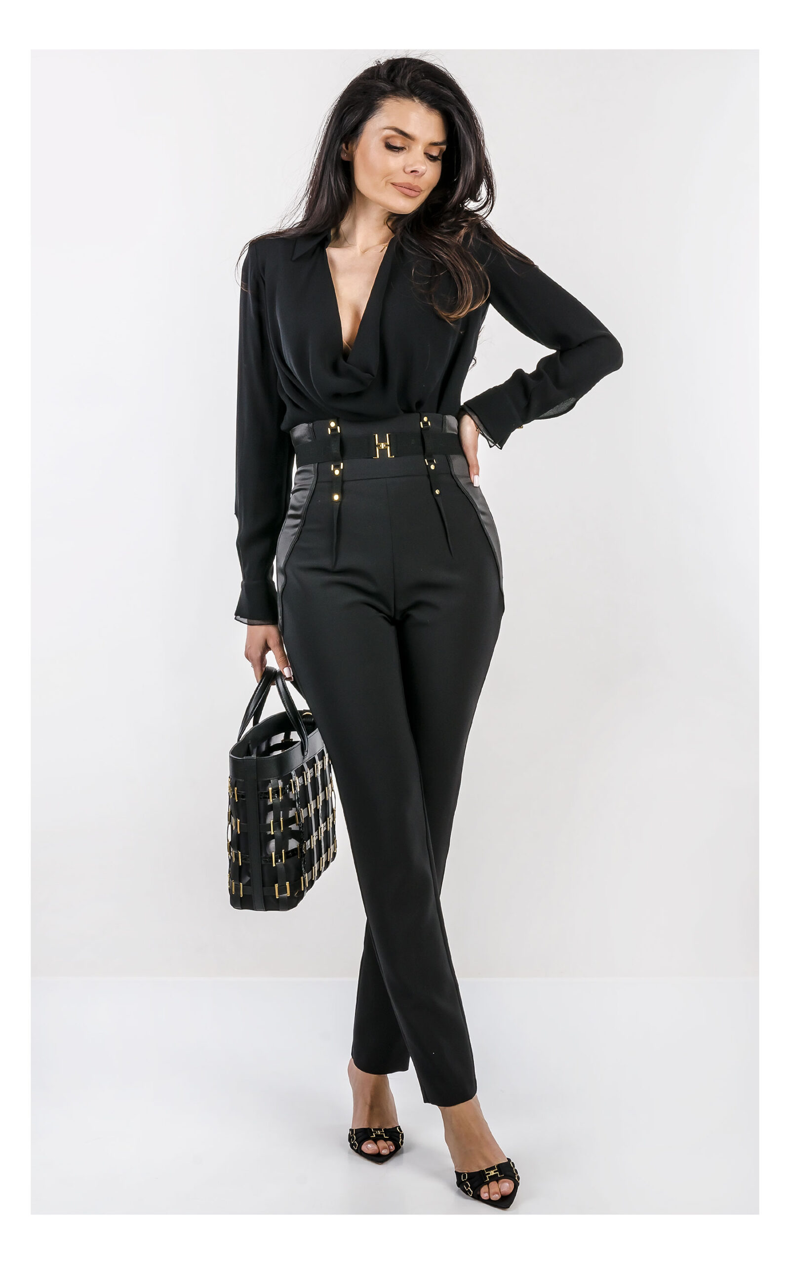 Na zdjęciu znajduje się modelka na jasnym, studyjnym tle, ubrana w elegancką stylizację w całości w kolorze czarnym. Ma na sobie czarne body z długim rękawem i głębokim dekoltem oraz dopasowane, wysokie spodnie z ozdobnym pasem i złotymi detalami. W dłoni trzyma czarną torebkę Elisabetta Franchi z dekoracyjnymi elementami w kolorze złota. Stylizację uzupełniają czarne klapki na obcasie z metalicznymi zdobieniami. Cała stylizacja jest od marki Elisabetta Franchi.