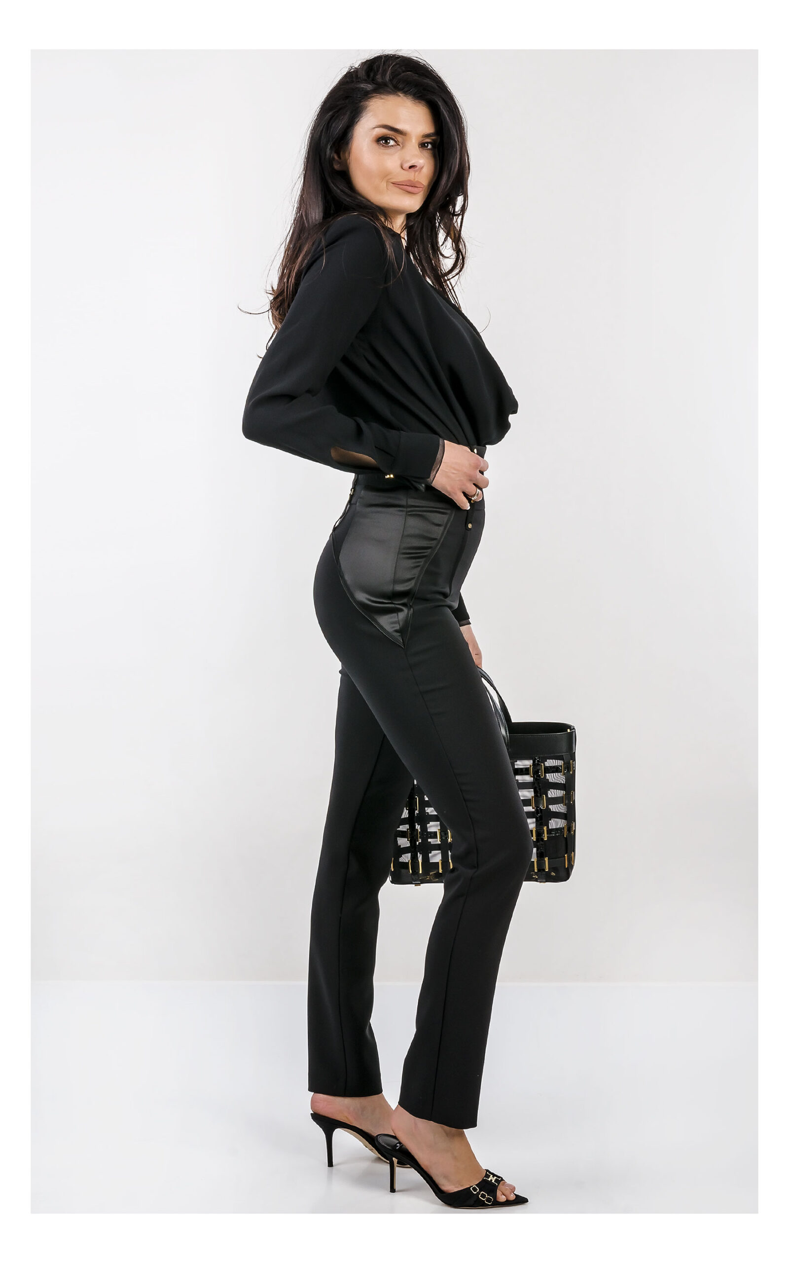 Na zdjęciu znajduje się modelka na jasnym, studyjnym tle, ubrana w elegancką stylizację w całości w kolorze czarnym. Ma na sobie czarne body z długim rękawem i głębokim dekoltem oraz dopasowane, wysokie spodnie z ozdobnym pasem i złotymi detalami. W dłoni trzyma czarną torebkę Elisabetta Franchi z dekoracyjnymi elementami w kolorze złota. Stylizację uzupełniają czarne klapki na obcasie z metalicznymi zdobieniami. Cała stylizacja jest od marki Elisabetta Franchi.Modelka stoi bokiem.